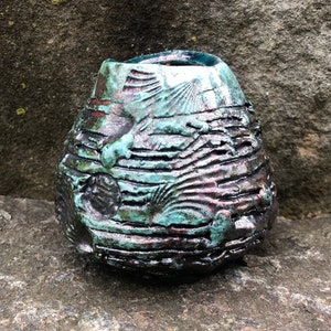 May include: A small, round ceramic vase with a textured, layered surface. The vase is a deep teal color with hints of brown and black.