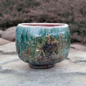 May include: A small, round ceramic bowl with a textured, crackled glaze in shades of green, blue, and copper. The bowl has a slightly uneven rim and a rounded base.