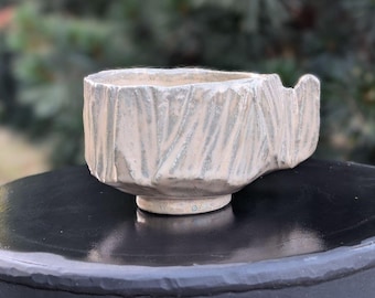 May include: A handmade ceramic cup with a textured, bark-like exterior. The cup is a light beige color with subtle blue-gray accents and a small handle. The cup sits on a small, round base.