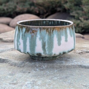 May include: A ceramic bowl with a white base and a turquoise and brown glaze. The glaze has a dripped effect, creating vertical streaks. The bowl has a rounded shape with a slightly flared rim and sits on a small, circular base.
