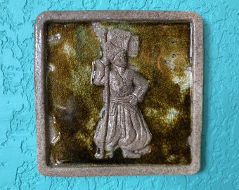 May include: A ceramic tile with a raised image of a person wearing a hat and holding a staff. The tile is brown with a green glaze and has a textured surface.