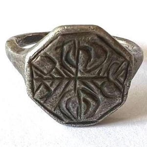 May include: A silver ring with an octagonal face featuring a detailed, engraved design. The design appears to be a stylized cross with four points.
