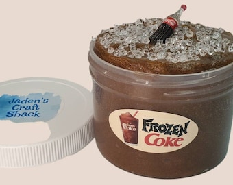 May include: A clear plastic jar filled with brown slime topped with white ice cubes and a miniature bottle of Coca-Cola. The jar has a white lid with the words "Jaden's Craft Shack" printed on it. The slime has a label that reads "Frozen Coke".