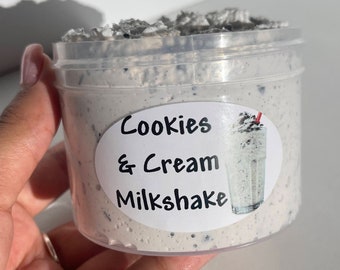 May include: A clear plastic container filled with a white and gray creamy substance. The container has a white label with the text "Cookies & Cream Milkshake" and a picture of a milkshake with a red straw.