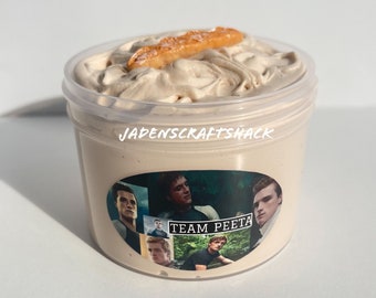 May include: A clear plastic container filled with light brown whipped cream topped with a brown cookie. The container has a sticker with the text "JADENSCRAFTSHACK" and a collage of images of a man with the text "TEAM PEETA".