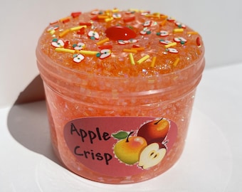 May include: A clear plastic container filled with orange-colored slime, decorated with apple-shaped sprinkles, red sprinkles, and a red cherry. The container has a label that reads "Apple Crisp" with apple illustrations.