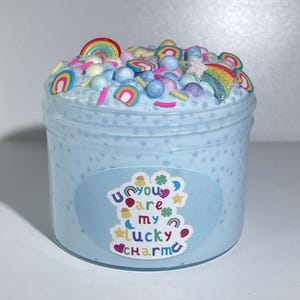 Op de afbeelding: Een lichtblauwe slime met regenboogsprinkels, gekleurde kralen en een sterbedel in een glazen pot. De pot heeft een etiket met de tekst "Jij bent mijn geluksbrenger".