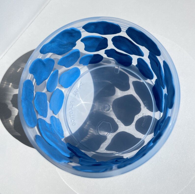 May include: A clear plastic cup with a blue cow print design. The cup is decorated with blue paint in a cow print pattern.