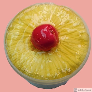 May include: A clear plastic container filled with a yellow pineapple-shaped slime with a red cherry on top.