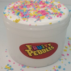 Op de afbeelding: Witte slijm met kleurrijke sprinkels in een doorzichtige plastic container met een rood label waarop "Fruity Pebbles" staat.