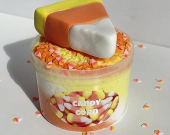 May include: A clear plastic container filled with yellow, orange, and white slime, decorated with candy corn-shaped sprinkles. A large candy corn sits atop the container. The label on the container reads "CANDY CORN".