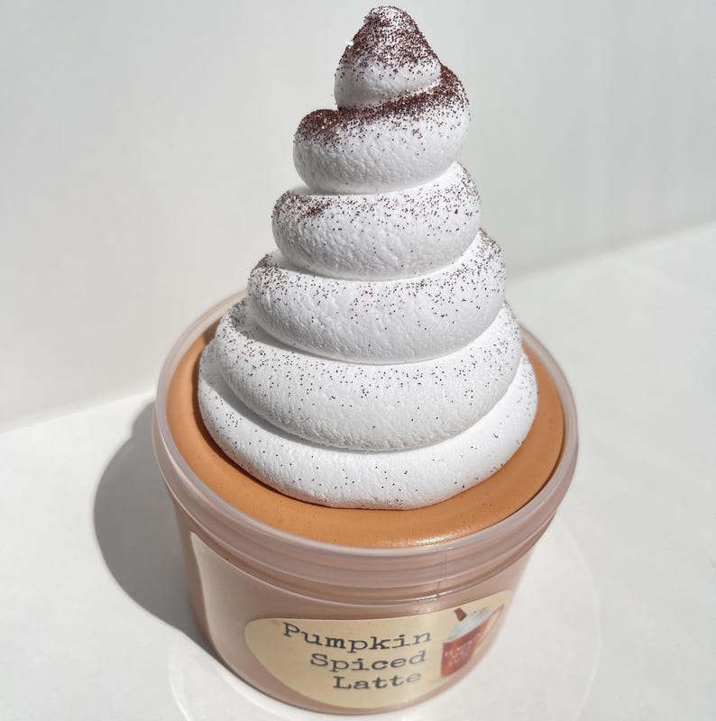 May include: A clear plastic container filled with a brown pumpkin spice latte slime topped with a large swirl of white whipped cream with brown sprinkles.