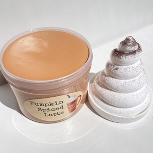 May include: A clear plastic container filled with a light brown slime labeled "Pumpkin Spiced Latte". Next to the container is a white, fluffy, whipped cream-like object with brown sprinkles.