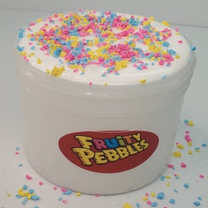 Op de afbeelding: Witte slijm met kleurrijke sprinkels in een doorzichtige plastic container met een rood label waarop "Fruity Pebbles" staat.