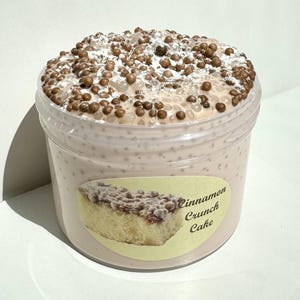 以下が含まれることがあります： 透明なプラスチック容器に、薄い茶色のスライムが入っており、茶色と白のトッピングがされています。容器には「シナモンクランチケーキ」というラベルが貼られています。