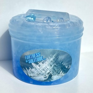 May include: A clear, blue container filled with a gel-like substance. The container has a clear lid with decorative blue crystals. An oval label on the front reads "Fortress of Solitude" with an image of an icy structure.