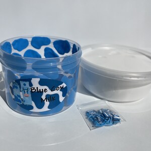 May include: A clear plastic container with a blue cow print design and the words "Blue Cow Milk" on a white label. The container is filled with white slime. A small plastic bag containing blue and white sprinkles is next to the container.