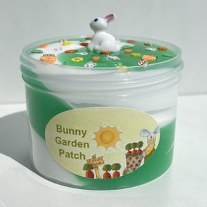 Puede incluir: Un slime verde y blanco con una figurita de conejo en la parte superior. El slime está en un recipiente de plástico transparente con una etiqueta que dice "Bunny Garden Patch".