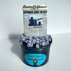Pode incluir: Um recipiente preto com um rótulo azul com as palavras "Daily Planet" e "Superman Saves the Day!". O recipiente é preenchido com contas de letras brancas e um item em forma de jornal com o título "Superman Saves the Day!"