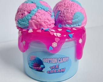 Puede incluir: Un recipiente de slime azul cubierto con dos bolas de helado de algodón de azúcar rosa y azul. El recipiente tiene una cobertura rosa goteante con chispas de corazón y flores. La etiqueta dice "Cotton Candy Ice Cream."