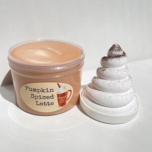 May include: A clear plastic container filled with a light brown slime labeled "Pumpkin Spiced Latte". A white whipped cream topping with brown sprinkles is next to the container.