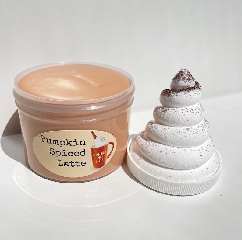 May include: A clear plastic container filled with a light brown slime labeled "Pumpkin Spiced Latte". A white whipped cream topping with brown sprinkles is next to the container.