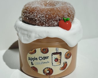 May include: A jar of brown slime topped with a sugared donut, white frosting, and a red apple charm. The jar has a label that reads "Apple Cider & Cider Donuts" with donut and coffee cup illustrations. The slime is a light brown color.