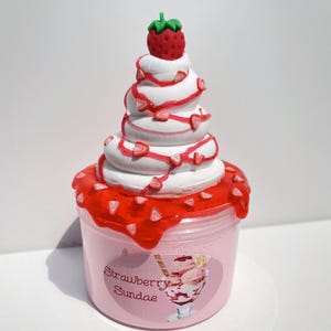 May include: A pink slime with a white whipped cream topping and red strawberry sauce. The slime is in a clear jar with a label that says "Strawberry Sundae".