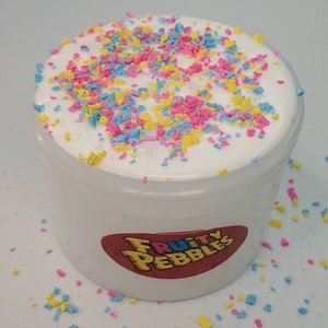 Op de afbeelding: Een witte slijm met roze, blauwe en gele sprinkles in een doorzichtige plastic container met een wit label waarop "Fruity Pebbles" staat.