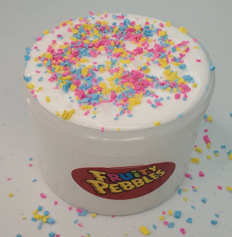 Op de afbeelding: Een witte slijm met roze, blauwe en gele sprinkles in een doorzichtige plastic container met een wit label waarop "Fruity Pebbles" staat.