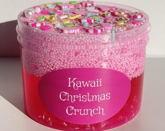 May include: A clear plastic pot filled with pink slime. The slime has a red base and is topped with pink foam beads and colourful sprinkles. A pink label reads "Kawaii Christmas Crunch."