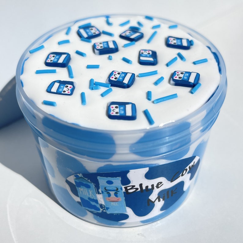 May include: A clear plastic container filled with white slime with blue sprinkles and blue milk carton charms. The container has a blue and white cow print design and the text "Blue Cow Milk".