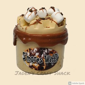May include: A brown and tan slime with a chocolate topping and marshmallows. The slime is in a clear container with a label that says "S'mores Waffle".
