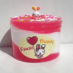 Pode incluir: Slime rosa e branco com um amuleto de coelho branco e granulado por cima. O slime está em um frasco transparente com um rótulo que diz "Kawaii Bunny".
