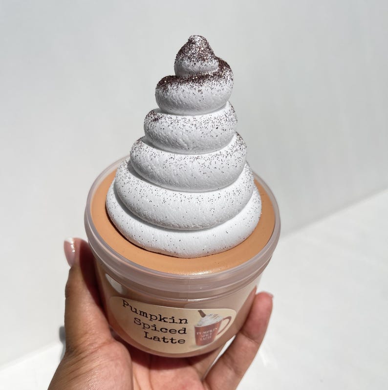 May include: A clear plastic container filled with a brown pumpkin spice latte slime with a white whipped cream topping with brown sprinkles on top. The container has a label that says "Pumpkin Spiced Latte".