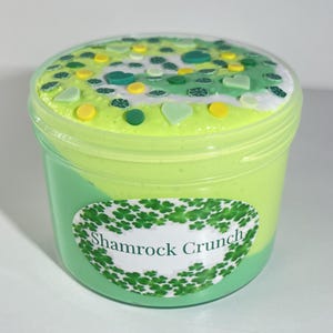 May include: A green and white slime in a clear plastic jar with a label that says "Shamrock Crunch". The slime is topped with green and white sprinkles.
