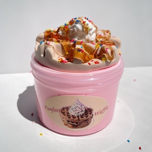 Op de afbeelding: Roze slijm in een doorzichtige container met een witte slagroom en een karamelsiroop eroverheen. De slagroom is versierd met kleurrijke sprinkels. De container heeft een label met de tekst "Funfetti Waffle".
