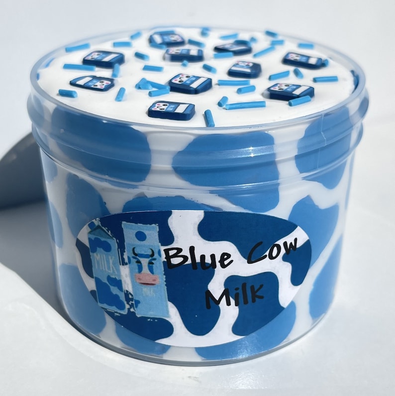 May include: A clear plastic container of blue slime with a white cow print design. The slime is topped with blue and white sprinkles and miniature blue milk cartons. A white label on the container reads "Blue Cow Milk".