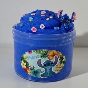 May include: A blue slime container with a Stitch character figurine on top. The slime is decorated with small pink and white flower-shaped embellishments. The container has a label with the word "Ohana" and a Stitch graphic.