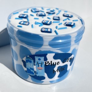 May include: A blue and white container of slime with a cow print design and the text "Blue Cow Milk". The slime is topped with blue and white sprinkles and small blue milk carton charms.