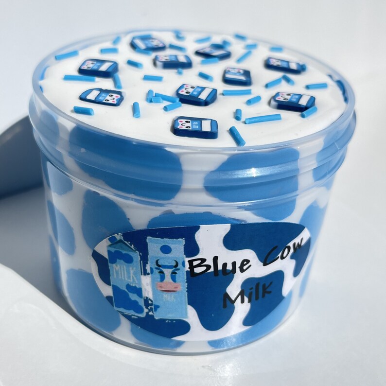 May include: A blue and white container of slime with a cow print design and the text "Blue Cow Milk". The slime is topped with blue and white sprinkles and small blue milk carton charms.