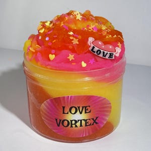 May include: A clear plastic container filled with colorful slime. The slime is layered with yellow, orange, and pink hues, topped with glitter and star-shaped confetti. A small heart-shaped charm with the word "LOVE" is attached. The container has the words "LOVE VORTEX" on the front.