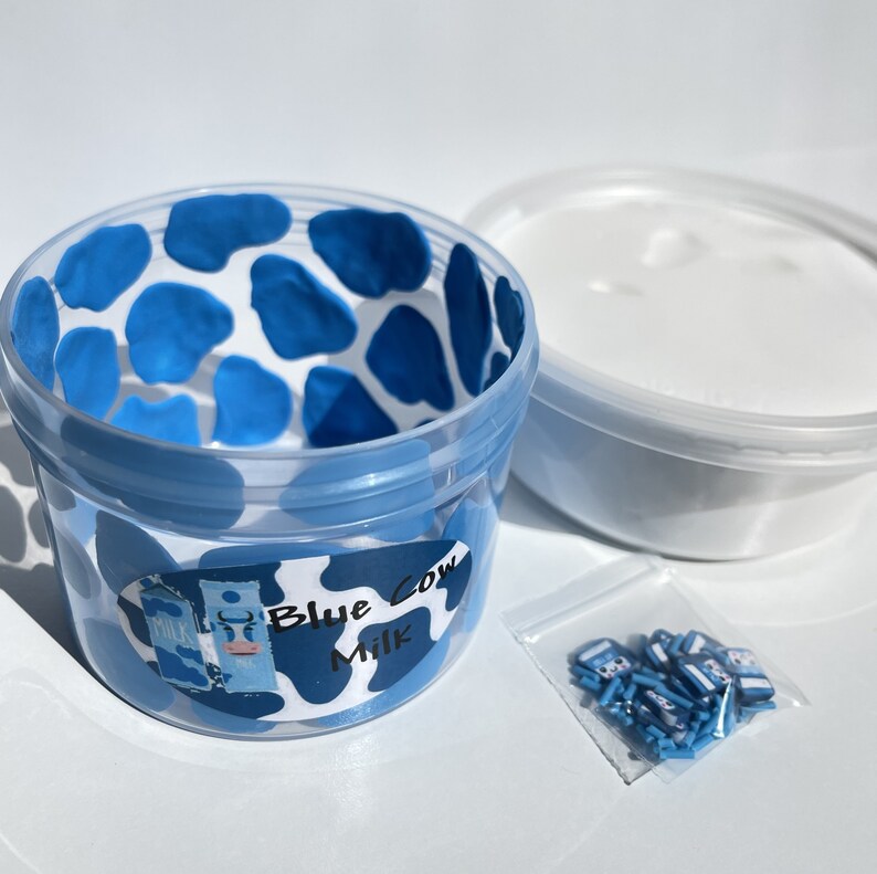 May include: A clear plastic container with a blue cow print design. The container is filled with white slime and has a label that reads "Blue Cow Milk". A small bag of blue cow-shaped charms is also included.