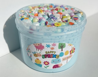 May include: A clear plastic container filled with light blue slime with colorful Easter-themed sprinkles and beads. The container has a label that says "Happy Spring" with a cute cartoon design of a bee, a sheep, a ladybug, and a flower.