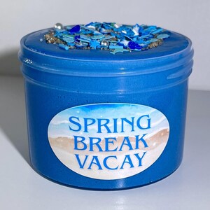 Pode incluir: Um recipiente azul de slime com um topo decorativo. O topo é coberto com glitter azul e prateado e confetes em forma de estrela. O recipiente tem um rótulo que diz "SPRING BREAK VACAY" com uma cena de praia.