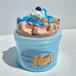 Op de afbeelding: Een blauwe plastic container met een wit deksel gevuld met lichtblauwe slijm. De slijm is bedekt met een wafeltjesvormige bruine en roze slijm met blauwe en paarse sprinkles. De container heeft een label met de tekst "Blueberry Waffles".