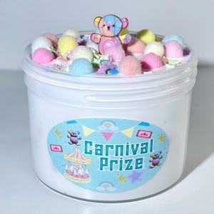 May include: A white container of slime with colorful pom-poms, sprinkles, and a small teddy bear figurine on top. The container has a label that reads "Carnival Prize" with a carousel illustration and teddy bear graphics.