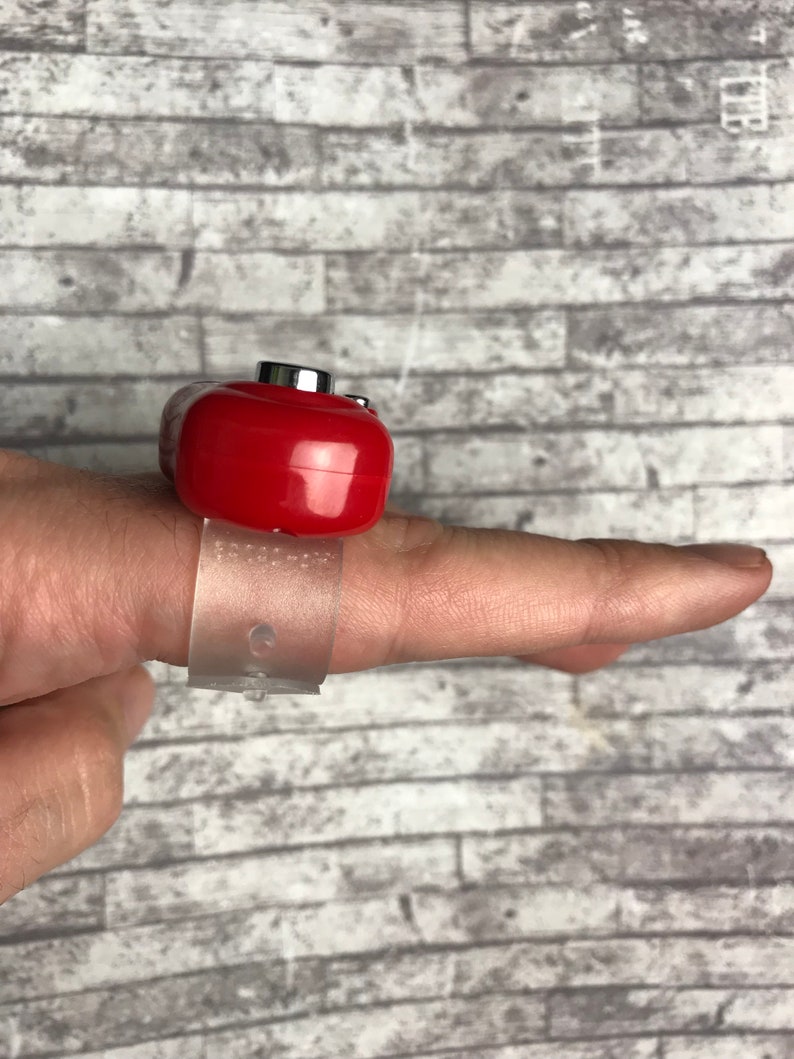 May include: A red ring with a clear plastic band. The ring has a small button on top.