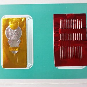 K&ouml;nnte beinhalten: Zwei Packungen N&auml;hnadeln, eine mit einer goldenen Folienverpackung und einem silbernen m&uuml;nzf&ouml;rmigen Design, und die andere mit einer roten Folienverpackung.