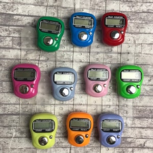 May include: Ten colorful digital tally counters with a clear plastic wrist strap. Each counter has a button labeled "COUNT" and "RESET".